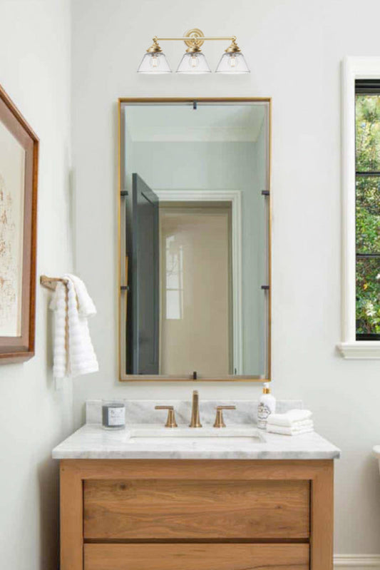 Triple lamp wall sconce in gold/brass with clear shades over a mirror in a classic bathroom. 