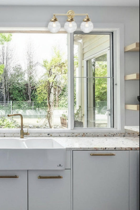 Two, 3 lampholder wall sconce in gold/brass with clear reeded glass ball shades over a white kitchen space with a windowed back splash. 