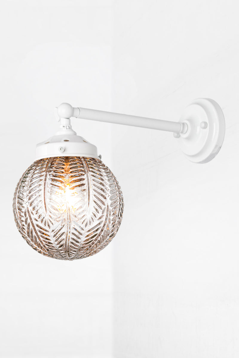 Long white, straight arm wall sconce with tiltable lampholder paired with a clear glass orb shade with intricate detailing. 