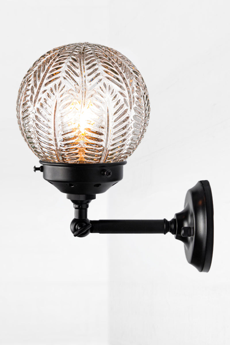 Short straight arm wall sconce with tiltable lampholder in black paired with a clear glass orb shade with intricate detailing. 