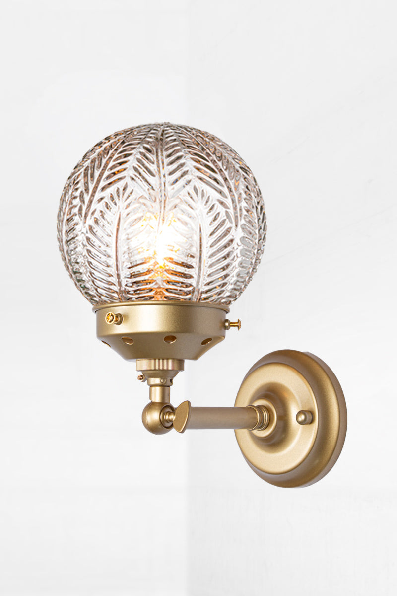 Short straight arm wall sconce with tiltable lampholder in gold/brass paired with a clear glass orb shade with intricate detailing. 