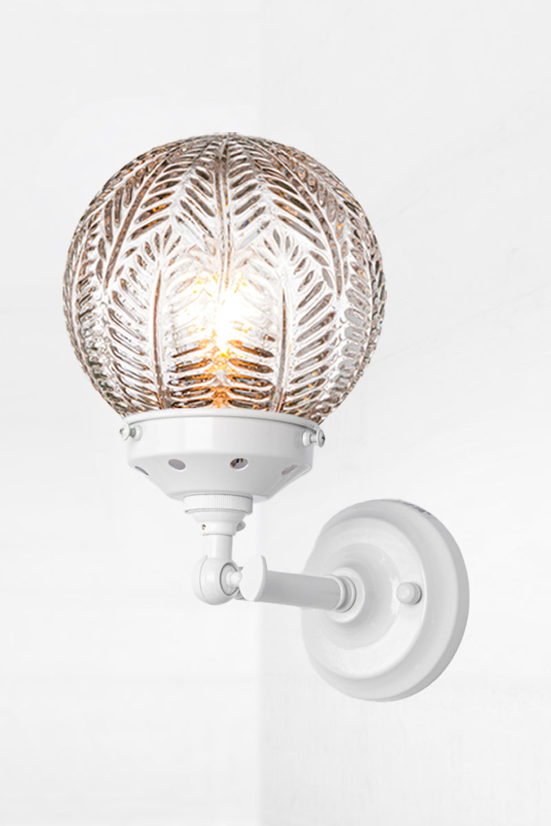 Short white, straight arm wall sconce with tiltable lampholder, paired with a clear glass orb shade with intricate detailing. 