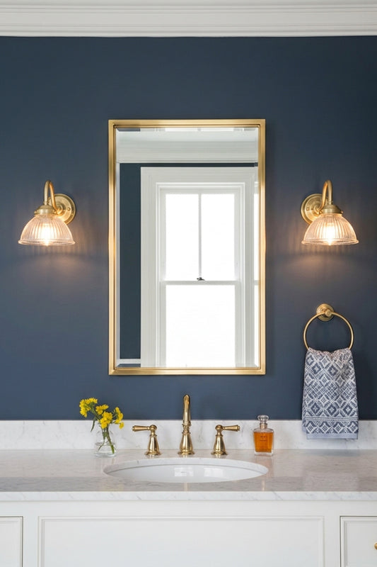 A framed wall mirror with a gold frame hangs on a dark navy blue wall above a white marble bathroom vanity. Two matching gold brass gooseneck wall lights with ribed glass light the space.