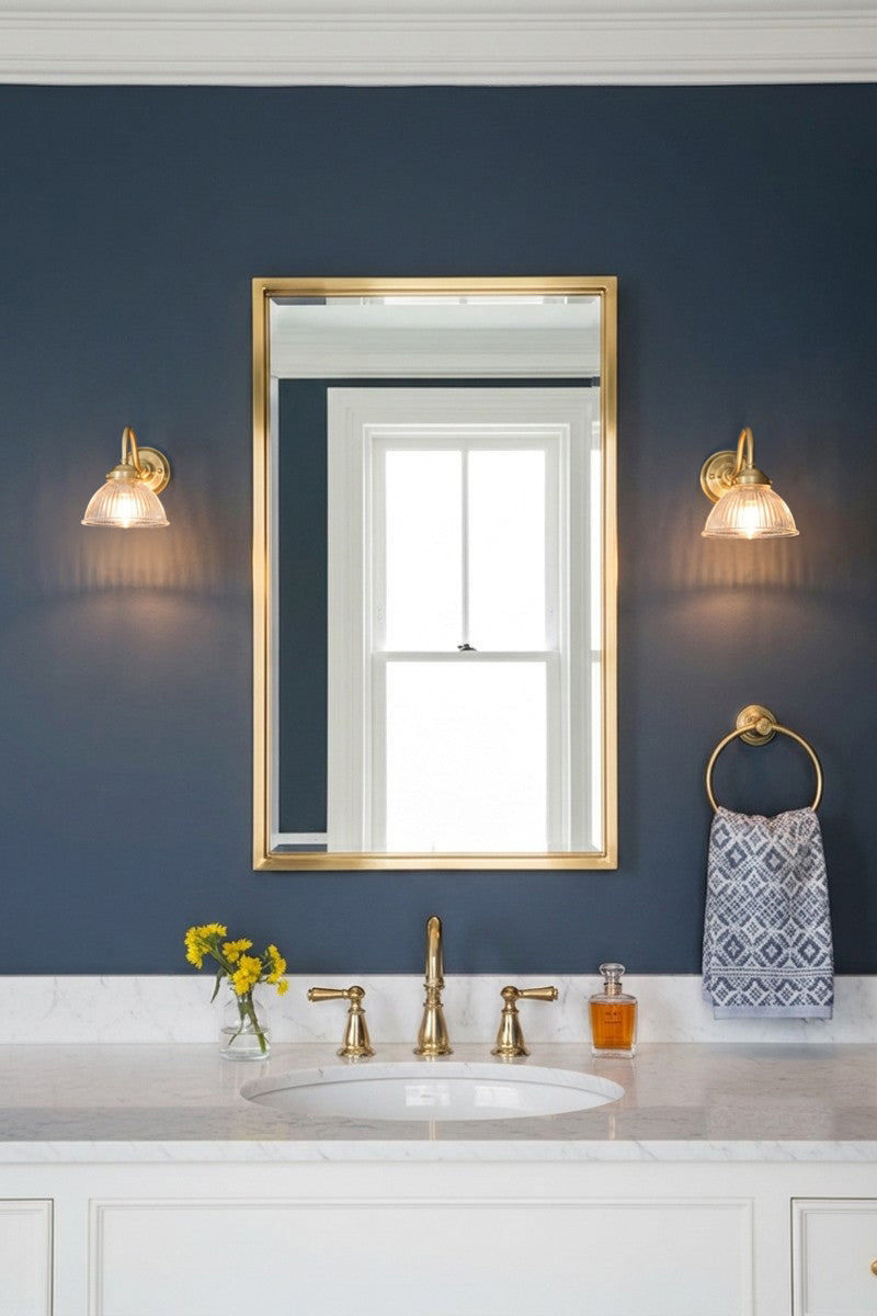 A framed wall mirror with a gold frame hangs on a dark navy blue wall above a white marble bathroom vanity. Two matching gold brass gooseneck wall lights with ribed glass light the space.