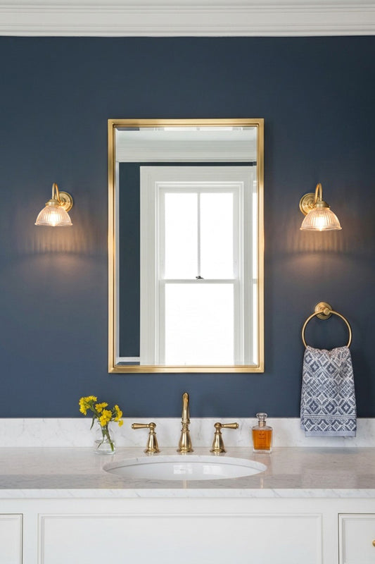 A framed wall mirror with a gold frame hangs on a dark navy blue wall above a white marble bathroom vanity. Two matching gold brass gooseneck wall lights with ribed glass light the space.