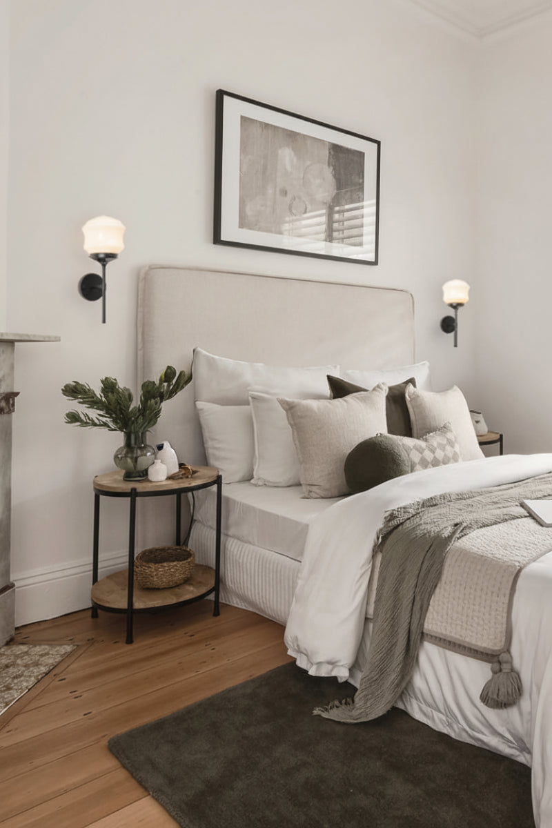Two black stemwall sconces with opal schoolhouse style shades as bedside lights in a room with grey and white soft furnishings.