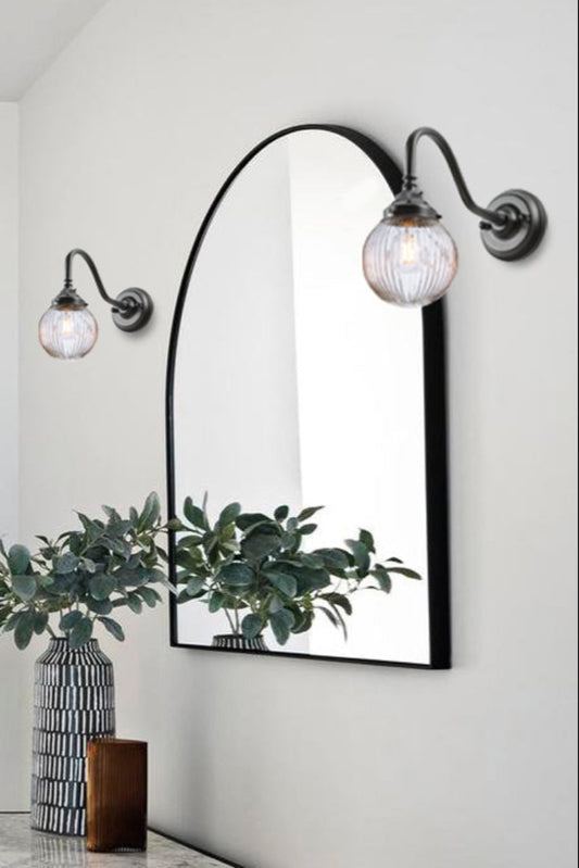 Two black gooseneck wall sconces with a clear reeded glass ball shades either side of an entryway mirror. 
