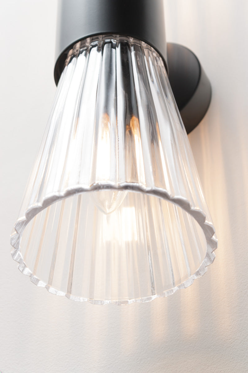 A modern wall sconce with a clear ribbed glass shade paired with 2 clear ribbed glass shades and sculptural steel body in black.