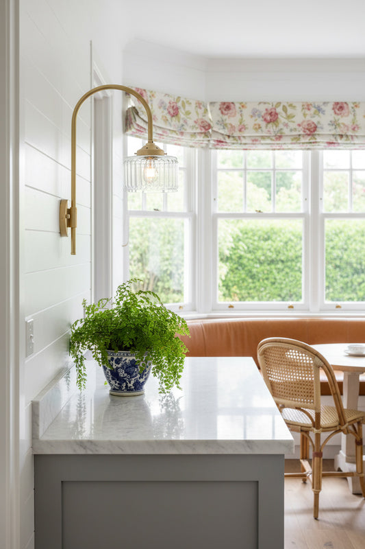Gold/brass shepherds hook wall light with a clear glass shade, over a side bench in a bright breakfast nook.