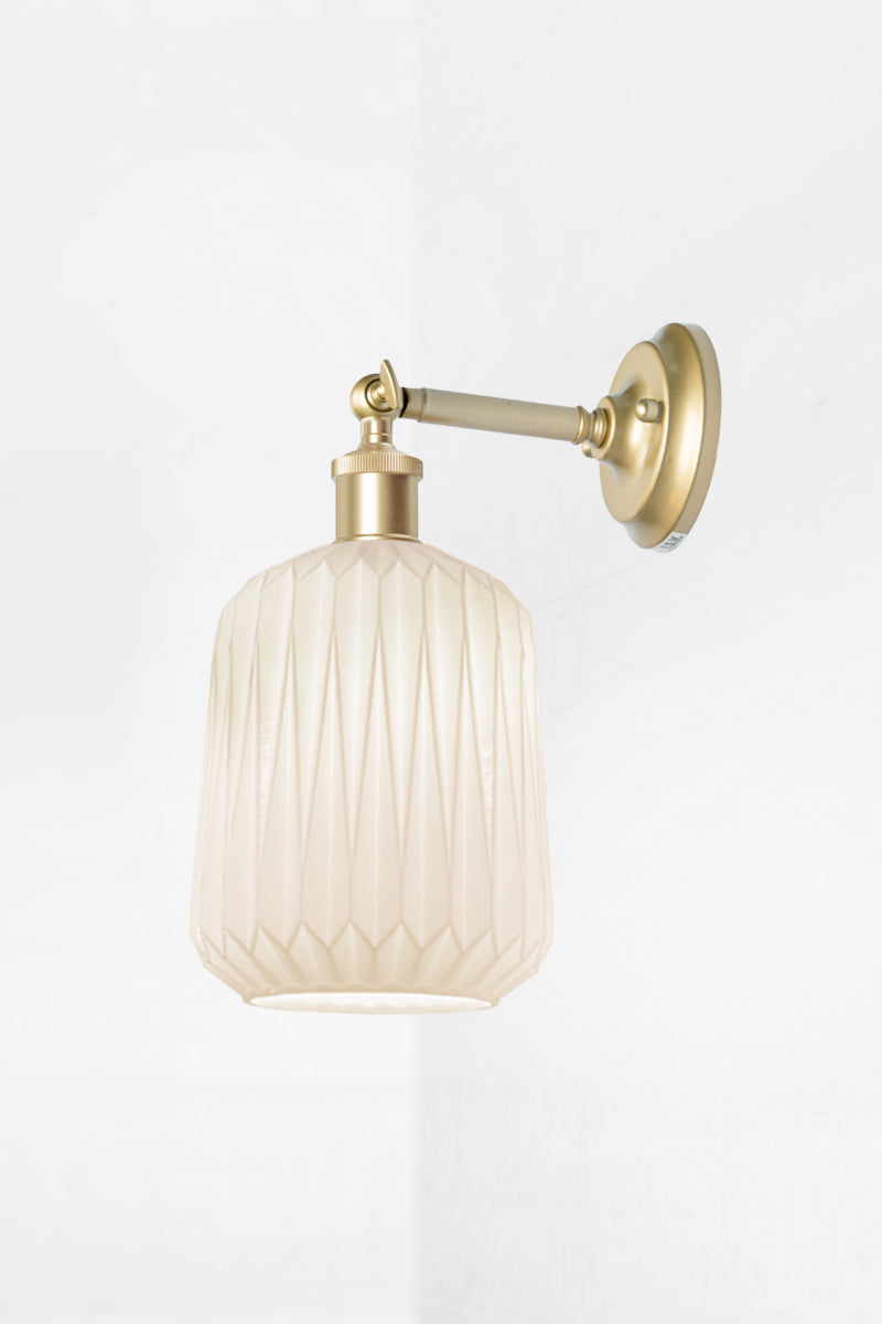 Wall sconce with a short gold/brass  straight arm tilted out holding an opal glass shade with geometric detailing. 
