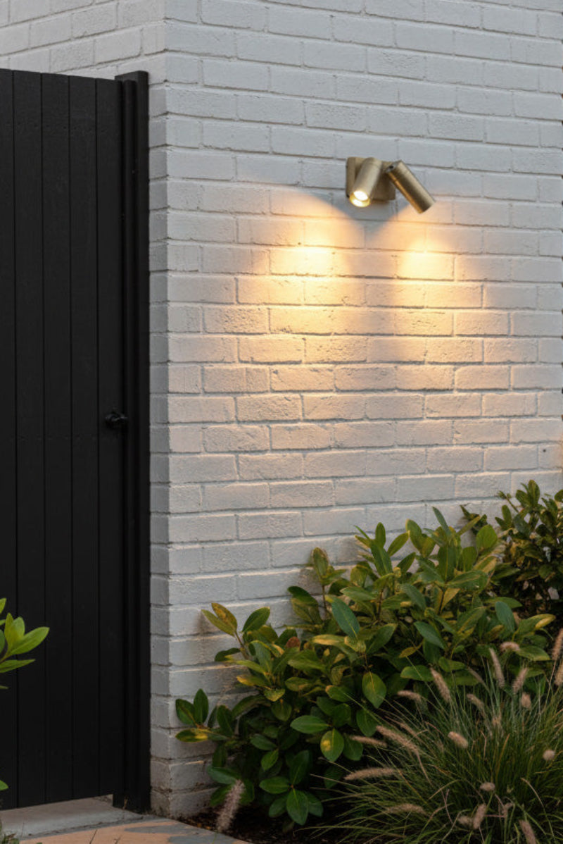 Modern outdoor brass wall light with two spotlight heads on a gray brick wall with garden plants.