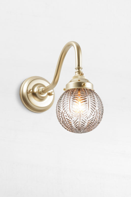Gooseneck arm wall sconce in gold/brass with a clear glass orb shade with intricate detailing. 