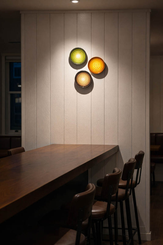 Modern wall lights with sage, amber, and grey shades on a white paneled wall above a wooden table. W1323-MIX-B-bar
