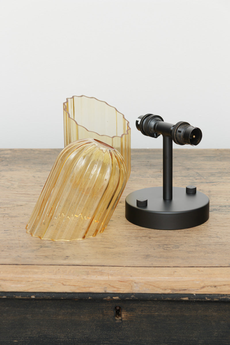 Black double wall sconce with two clear glass shades with fluted detailing and angled cut design place on a timber bench. 