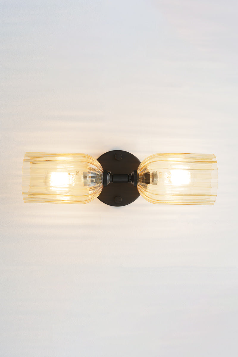 Black double wall sconce with two elongated amber glass shades with fluted detailing installed horizontally. 