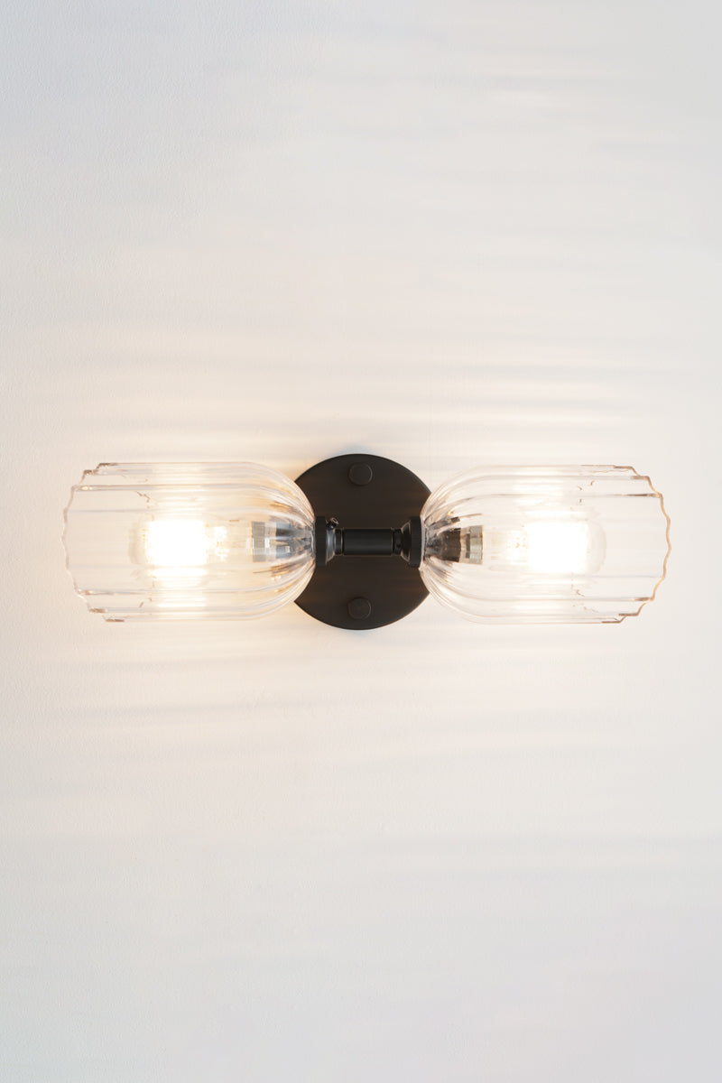 Black double wall sconce with two elongated clear glass shades with fluted detailing installed horizontally. 