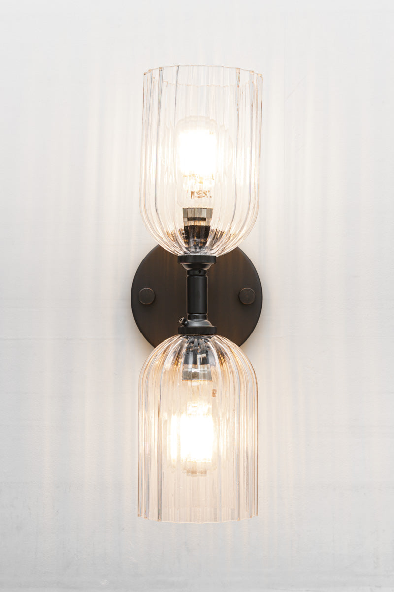 Black double wall sconce with two elongated clear glass shades with fluted detailing.