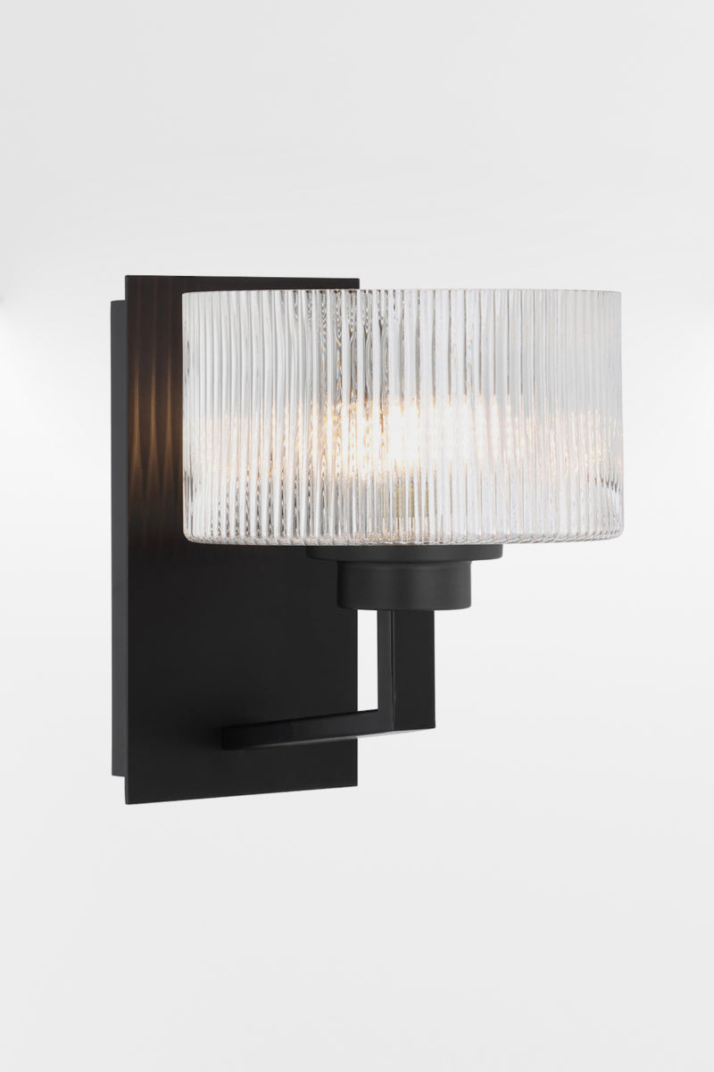 Wall sconce in black with a rectangular mount plate and arm, holding a clear reeded cylindrical glass shade.