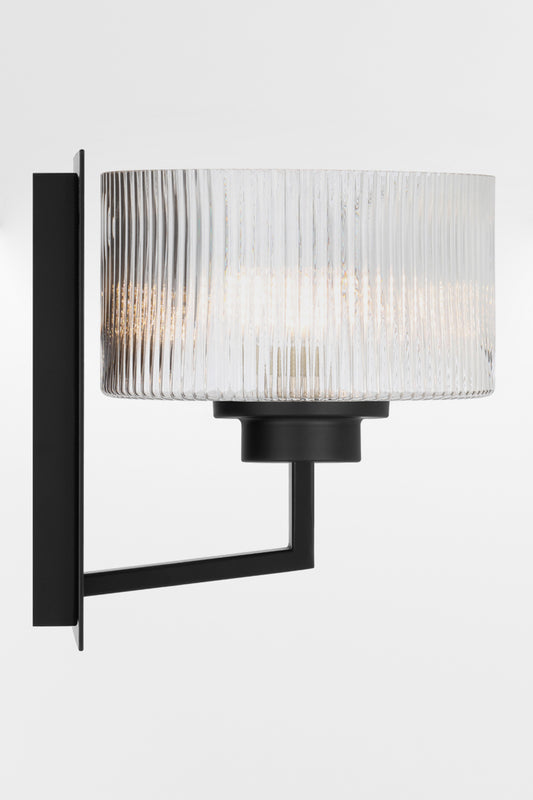Side view of the wall sconce in black with a rectangular mount plate and arm, holding a clear reeded cylindrical glass shade.