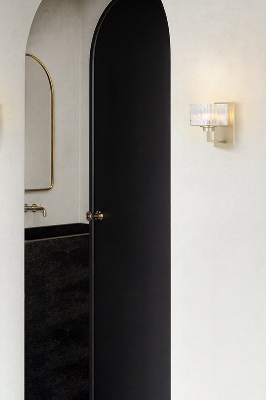 Antique Silver wall sconce with a reeded glass shade as hall lighting next to an arched doorway od a white, black and gold bathroom space. 