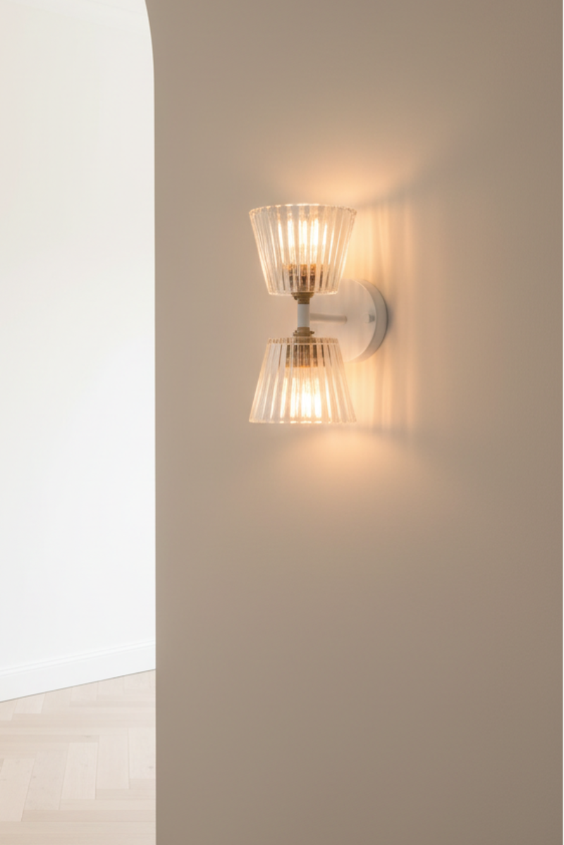 A modern wall-mounted sconce with two ribbed glass shades and a brushed metal base, glowing warm light on a beige wall next to an arched doorway.