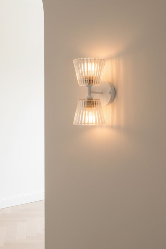 A modern wall-mounted sconce with two ribbed glass shades and a brushed metal base, glowing warm light on a beige wall next to an arched doorway.