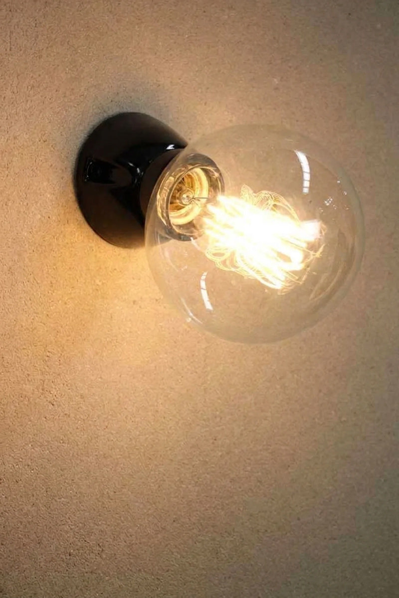 Delmont black glazed ceramic wall mount with a lit clear globe filament bulb, on a textured grey wall