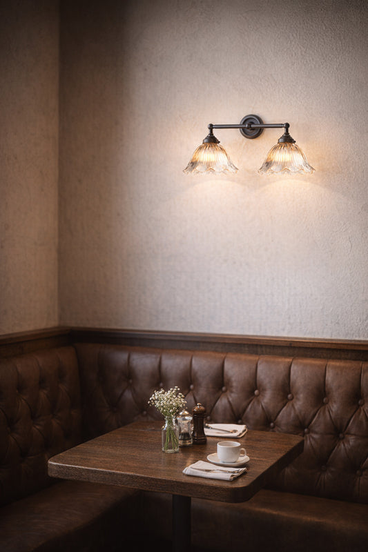 Double swivel wall sconce in black with 2 clear reeded glass shades with frilled edges over a brown leather dining booth. 