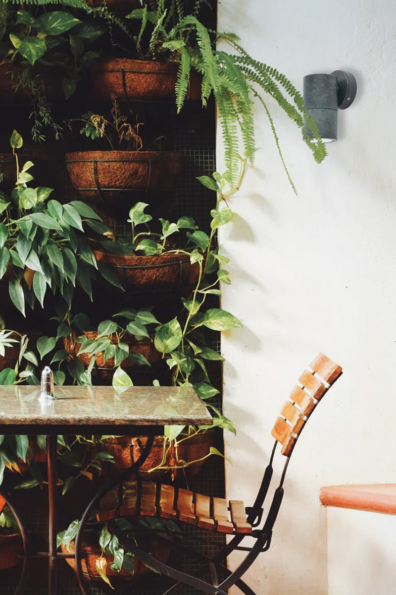 Solid brass fixed wall spotlight wih a green patina finish on a white outdoor wall with a sitting area and greenery. 