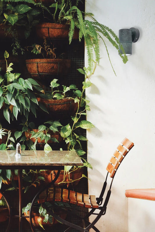 Solid brass fixed wall spotlight wih a green patina finish on a white outdoor wall with a sitting area and greenery. 