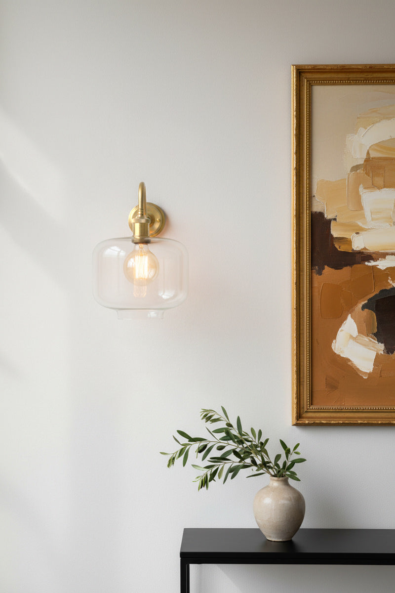 A brass wall sconce with a clear glass shade and visible filament bulb, mounted on a white wall in a dining room interior setting with a framed abstract painting 