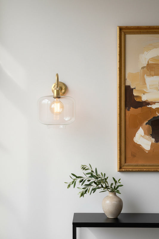 A brass wall sconce with a clear glass shade and visible filament bulb, mounted on a white wall in a dining room interior setting with a framed abstract painting 