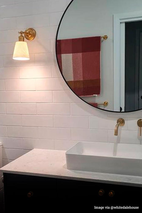 Gold/brass wall light with small ceramic shade next to a mirror in a bathroom.