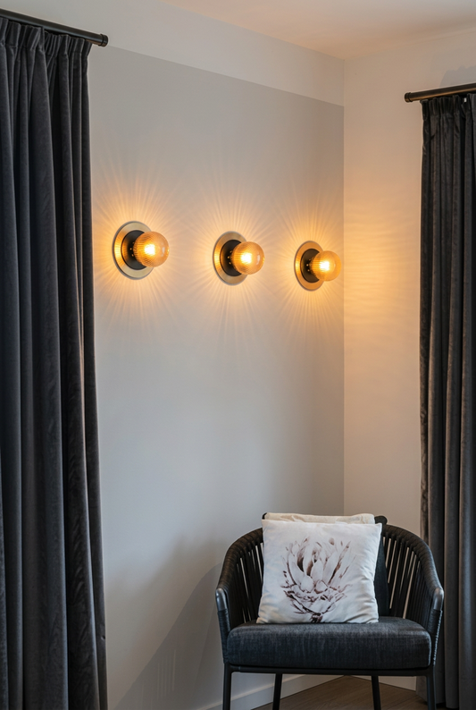 Three modern wall sconces dark amber reeded glass ball light with a small black disc and a large brass disc mounted on a light gray wall, flanked by dark gray curtains, with a dark gray armchair and a white decorative pillow in the foreground.