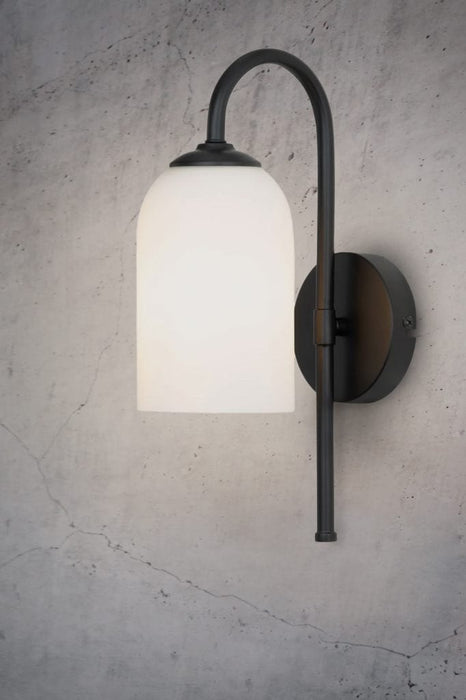 Wall light with cylindrical opal glass shade and black metalware build affixed on textured wall from a different angle. 