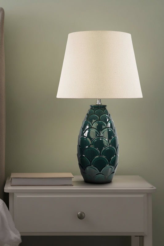 Table lamp with dark green scale-like base and shade in an ivory finish on a bedside table.
